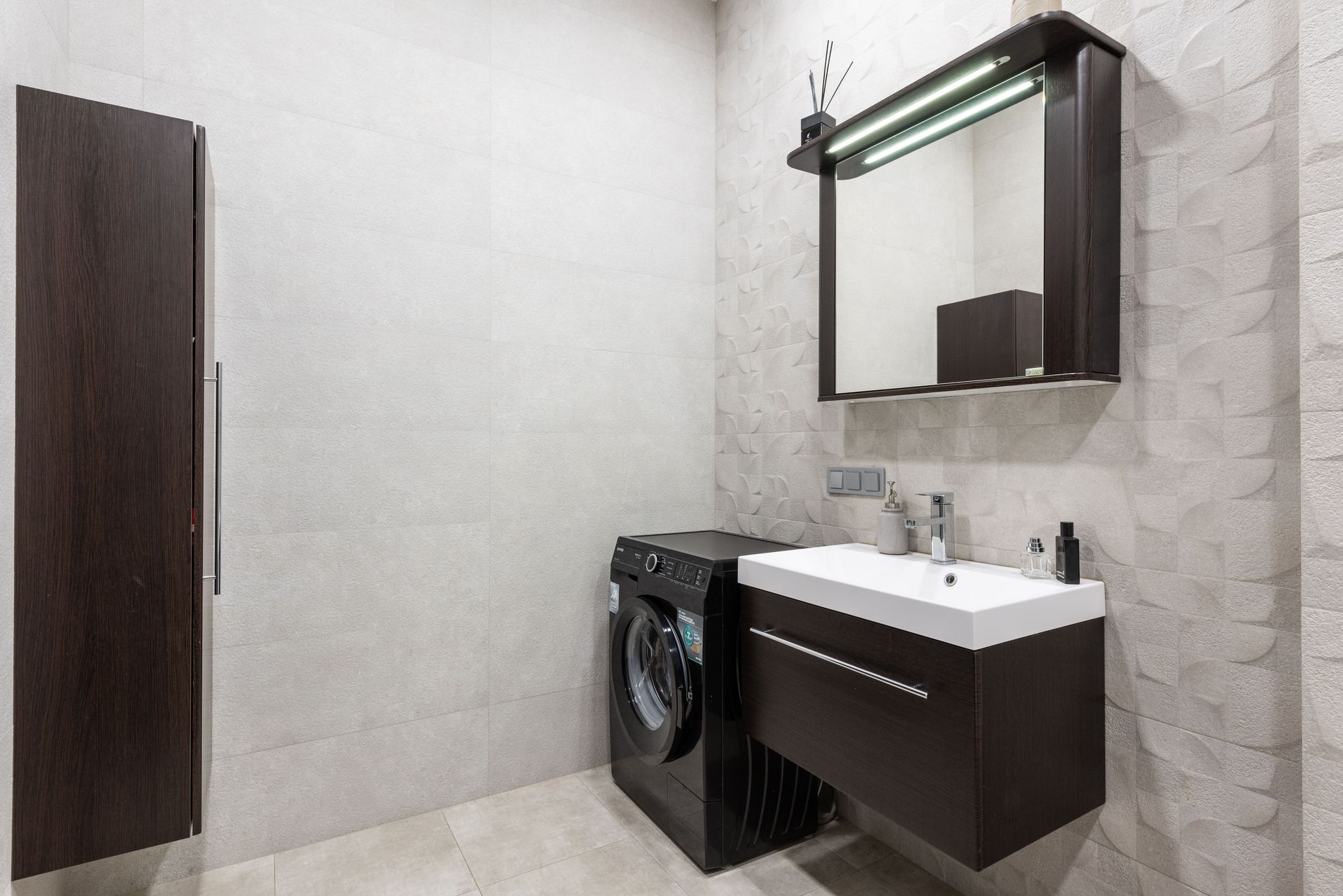 Modern black washing machine placed at wall near sink with dispenser and mirror in stylish light bathroom with brown cupboard